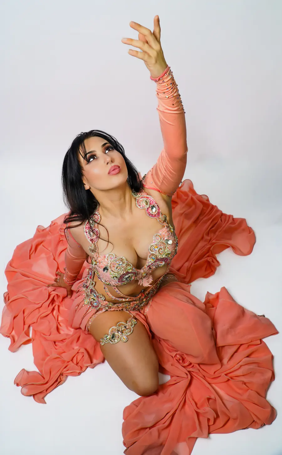 Belly dancer Azeeria Azizah kneeling in a coral costume, one arm reaching upward