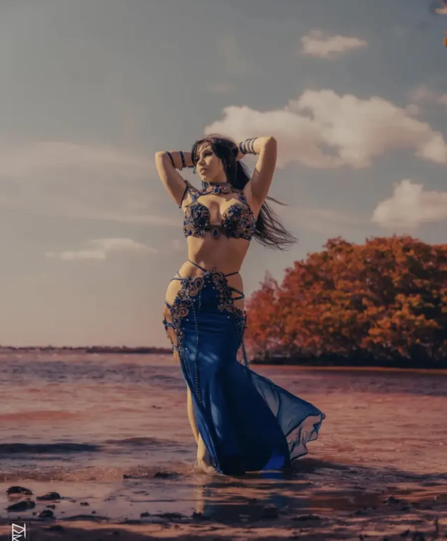 Belly dancer Azeeria Azizah standing in the water at sunset in a blue and gold costume
