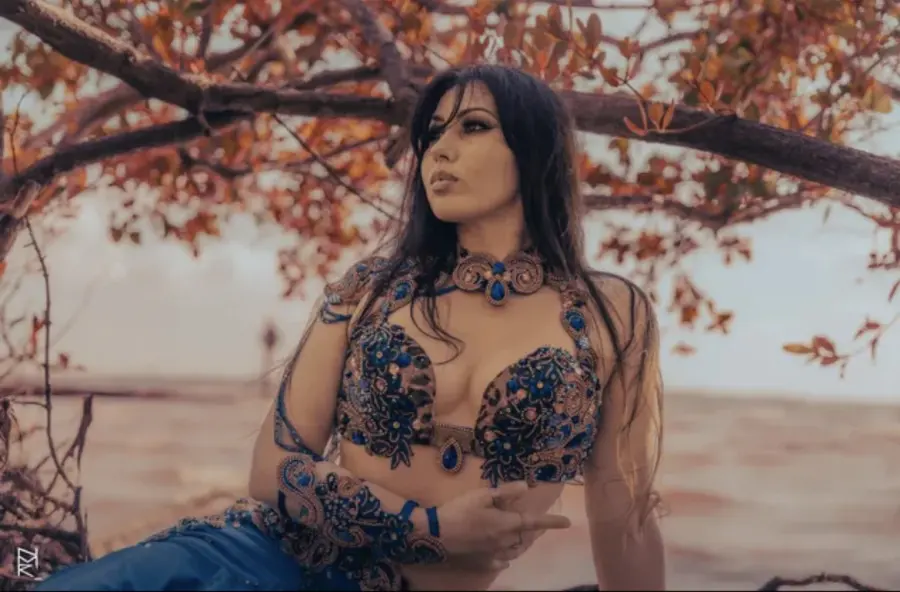 Belly dancer Azeeria Azizah seated at the waterline beneath autumn branches, wearing a jeweled collar